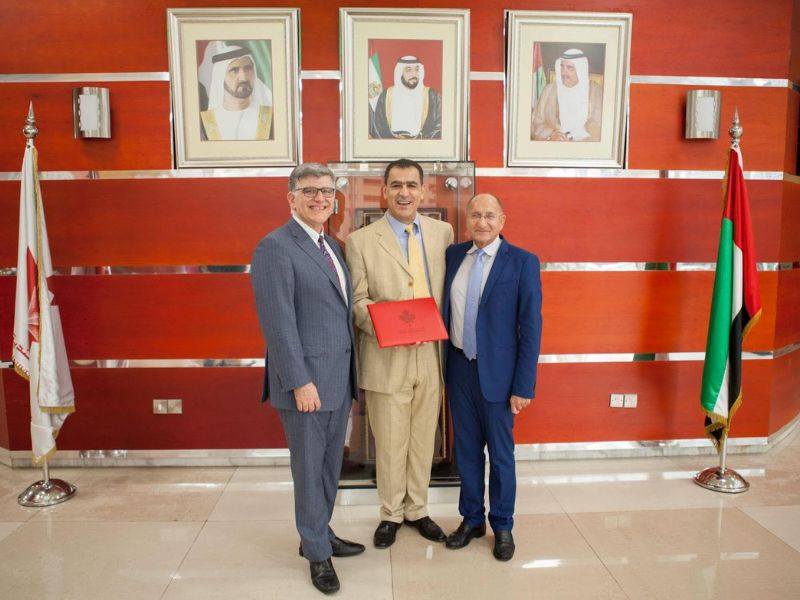 Three individuals in business attire stand together and pose, with one holding a certificate. Portraits and the Canadian and United Arab Emirates flags are displayed in the background.